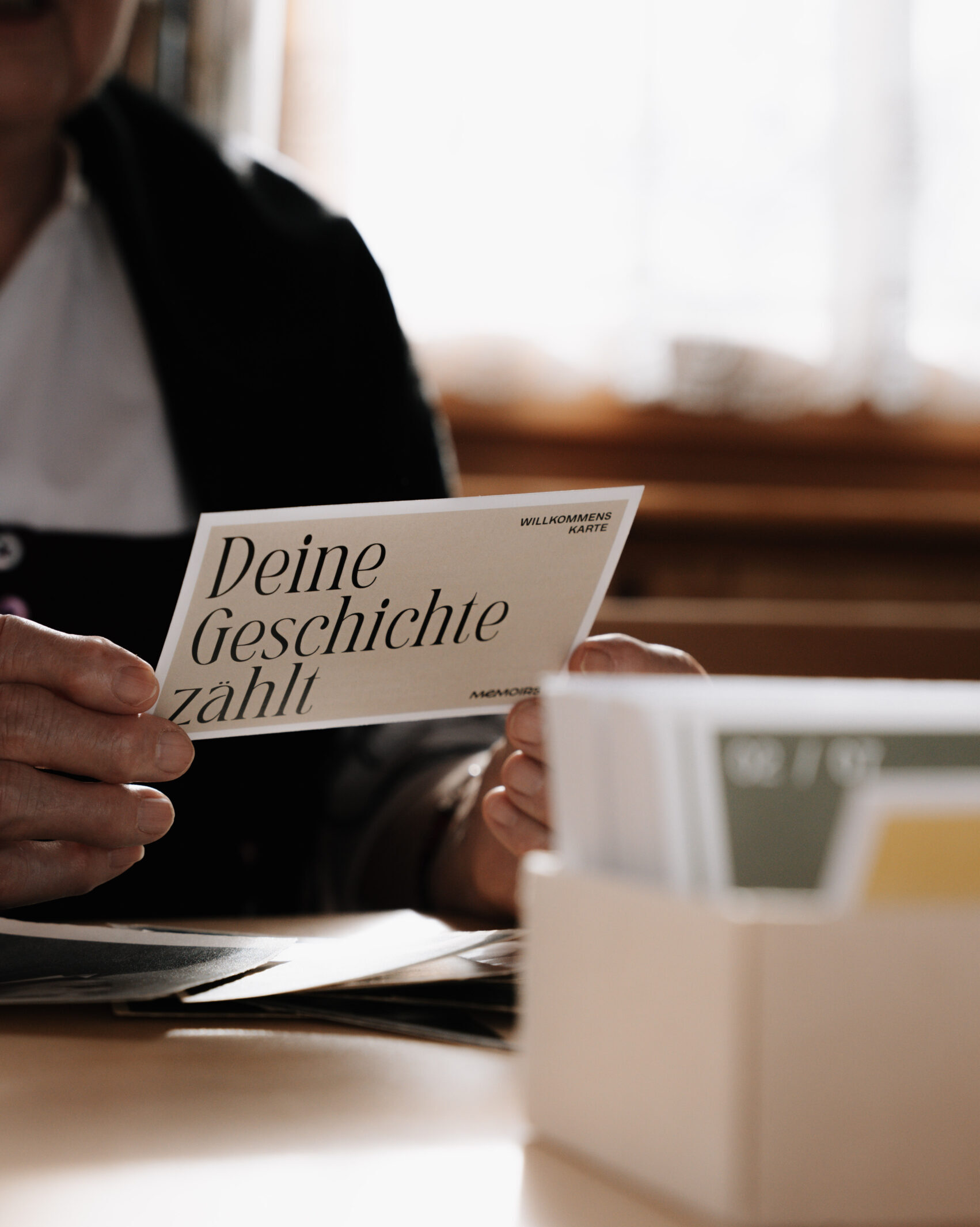 Hand hält ein Memoirs-Kartenset in der Hand, Deine Geschichte zählt, im Hintergrund Oma zu sehen, jetzt erhältlich im Memoirs Shop